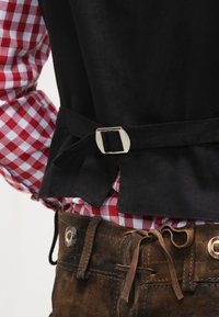 Black vest with a silver buckle detail, worn over a red and white checkered shirt. Featured on brown textured pants with a tie closure.