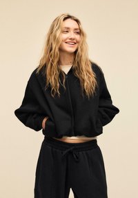 Črna zip-up kratka jopa z ohlapnim krojem in širokimi rokavi. Skupaj s temnimi trenirkami, oba izdelana iz mehkega materiala.