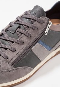 Baskets grises en daim et cuir, à lacets avec une fermeture éclair, détail bleu, et semelle plate de couleur claire.