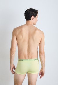 Moške bokserice svetlo rumene barve z oprijetim krojem in črnim logotipom Calvin Klein na pasu. Zadnji pogled.