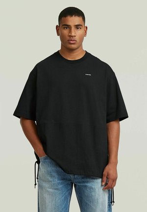 Giovane uomo che indossa una maglietta nera a maniche corte, larga, con un piccolo logo, e jeans azzurri, in piedi con una mano in tasca.