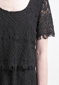 Svart spetsöverdel med vågkant, korta ärmar och intrikata blommönster. Texturen är detaljerad och lager på lager.