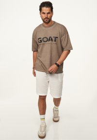 Braunes Oversize-T-Shirt mit schwarzem "GOAT"-Schriftzug, kombiniert mit weißen strukturierten Shorts und cremefarbenen Sneakern, dazu gestreifte Socken.