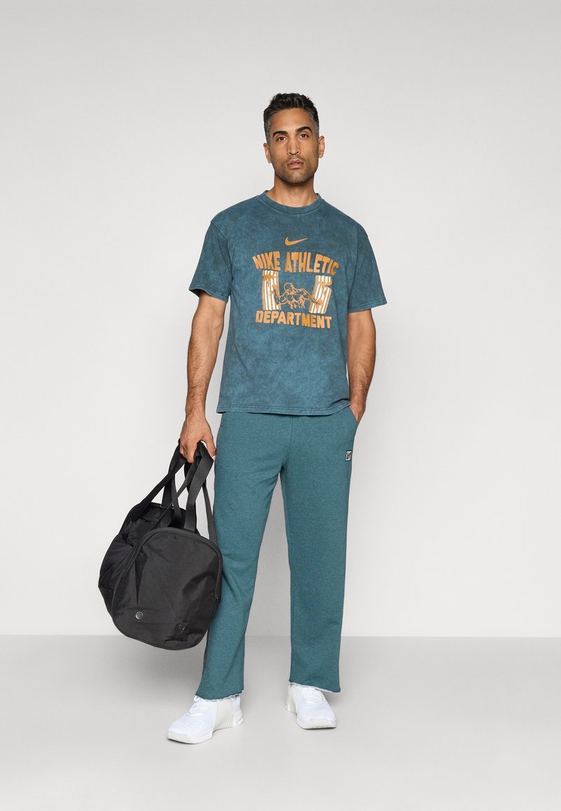 Sportssæt bestående af en teal grafisk t-shirt med orange tekst, matchende bukser og hvide sneakers, samt en sort gymnastiktaske.