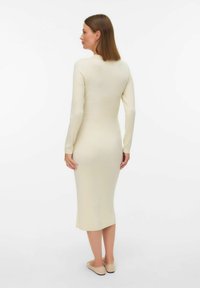 Krämfärgad ribbad midiklänning med långa ärmar och figurnära siluett, med rund halsringning och subtil struktur. Modellen bär beige ballerinaskor.