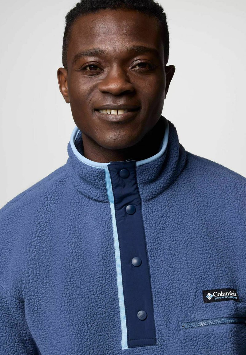 Polarowa bluza w kolorze niebieskim z teksturowanym materiałem, wysokim kołnierzem i kontrastowym panelem w kolorze granatowym. Posiada napy oraz kieszeń na zamek.