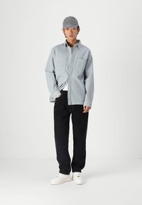 Lys blå skjorte med knapper og to frontlommer, kombinert med svarte bukser og hvite joggesko. Iført en grå caps, står på en ensfarget bakgrunn.