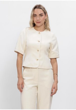 Kvinde med blondt hår iført cremefarvet kortærmet knapbluse og matchende højtaljede bukser, stående mod en ensfarvet hvid baggrund.