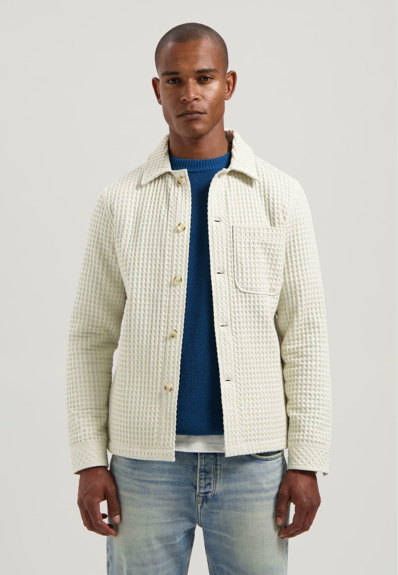 Giacca bianca a trama con colletto e tasca frontale, indossata sopra un maglione di maglia blu. I jeans in denim chiaro completano l'outfit.