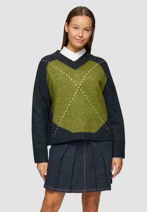Übergroßer V-Ausschnitt-Pullover in Dunkelgrün und Schwarz mit einem strukturierten, Rautenmuster. Kombiniert mit einem plissierten Jeansrock.