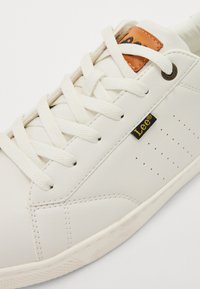 Valge nahast sneaker, millel on lamecid, perforeeritud detailid ja pruunist nahast silt. Tekstureeritud tallad ja peened õmbluse aktsendid.