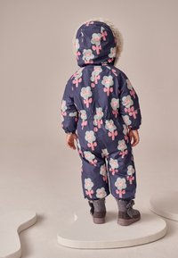 Combinezon de zăpadă albastru închis cu modele florale în roz și verde, cu glugă captusită cu blană artificială și manșete elastice, asortat cu cizme maro.