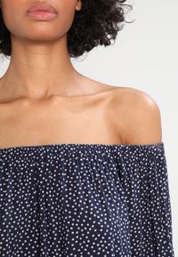 Kvinna med lockigt hår som bär en marinblå av-axel-top med vita prickar, visar nacke, axlar och övre bröst.