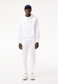 Vit zip-up hoodie och matchande byxor i mjukt material, bärs med en blå keps och vita sneakers med marinblå detaljer.