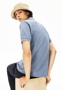 Blå pikétröja med korta ärmar, strukturerad tyg, bärs med marinblå byxor. Modellen bär en beige bucket hat, sittande på en trälåda.