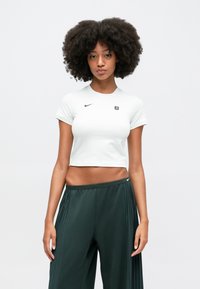 Balta īsām piedurknēm apgriezta t-krekls ar apaļu kakla izgriezumu un mazu melnu Nike logo; kombinēts ar tumši zaļiem krokotiem biksēm.