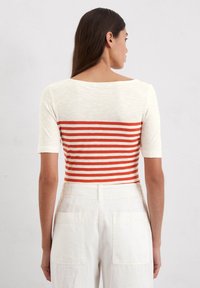 T-shirt met korte mouwen, met een crèmekleurige bovenkant en horizontale rood-oranje strepen aan de onderkant. Zachte stof, bootnek, en getailleerd ontwerp.