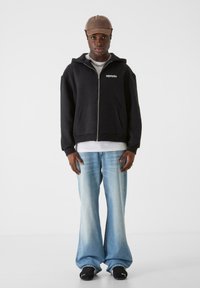 Svart hettejakke med glidelås og frontlommer, hvit grafisk logo; kombinert med lyseblå flagrende jeans og svarte sneakers. Beige caps på hodet.