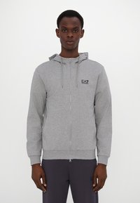 Siva zip-up dukserica izrađena od mekanog materijala, s kapuljačom, prednjim džepovima i suptilnim EA7 logom na prsima. Glatka tekstura.