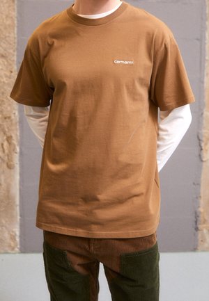 Osoba ubrana w brązowy, warstwowy t-shirt Carhartt z krótkim rękawem na białą koszulę z długim rękawem oraz spodnie z dwukolorowego sztruksu, stojąca w pomieszczeniu.