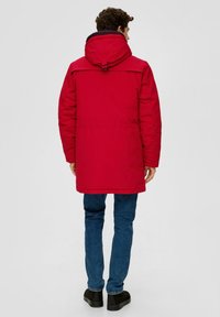 Giacca imbottita rossa con cappuccio, dotata di yoke posteriore, cinturino per cappuccio regolabile e taglio dritto; abbinata a jeans blu e scarpe nere.