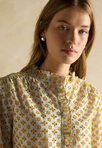 Cămașă albă cu broderie florală galbenă, guler cu volane și detaliu cu nasturi pe partea din față. Modelul poartă cercei argintii.