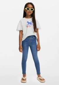 Bílý tričko s barevným potiskem tygra, modré legíny a žluté sluneční brýle. Má krátké rukávy a volný střih; sandály se zdobením ve tvaru srdce.