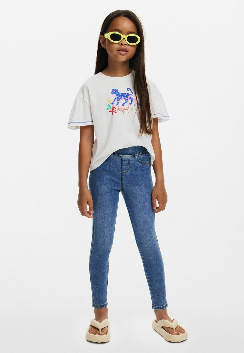 Bílý tričko s barevným potiskem tygra, modré legíny a žluté sluneční brýle. Má krátké rukávy a volný střih; sandály se zdobením ve tvaru srdce.