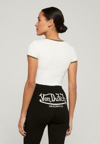 Bílé zkrácené tričko s zelenými akcenty v kombinaci s vysokými černými legínami, které mají vzadu bílé logo "Von Dutch Originals".