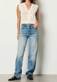 Háčkovaný pletený top bez rukávů v krémové barvě s výstřihem do V, kombinovaný s světle modr ými širokými džíny, nošený s černými mokasíny.