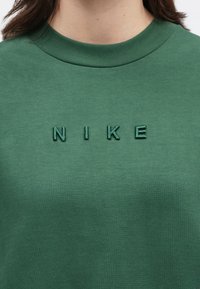 Πράσινο φούτερ με στρογγυλή λαιμόκοψη και φωτισμένο κεντημένο λογότυπο "NIKE" στο στήθος, το οποίο φ οριέται από άτομο με σκούρα καστανά μαλλιά.