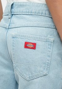 Τσέπη πίσω από τζιν σε ανοιχτό μπλε χρώμα με κόκκινο λογότυπο Dickies, φορεμένη από άτομο με εμφανές χέρι στη δεξιά πλευρά.