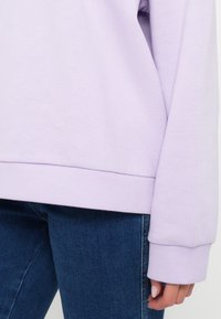 Personne portant un sweat-shirt violet clair à manches longues et un jean en denim bleu, montrant le bas du torse et le bras droit avec la main visible.
