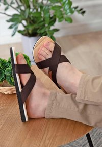 Brown cross-band sandals with a wooden sole, featuring a textured fabric strap and a distinct orange logo on the side.