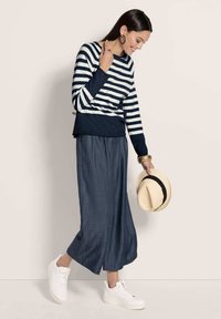 Frau in navy- und weißgestreiftem Langarmshirt, blauen weiten Hosen, weißen Sneakers, hält einen beigen Hut mit schwarzem Band, lächelt und schaut nach unten.