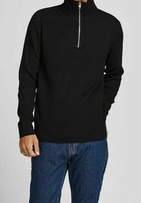 Homme portant un pull noir à manches longues côtelé avec une fermeture éclair à l'avant et un jean bleu, debout avec une main dans la poche.