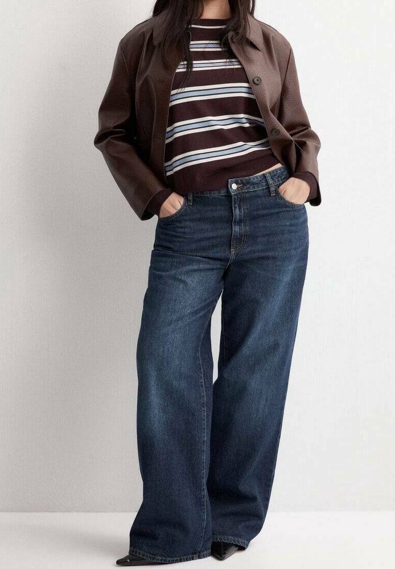Personne portant une veste en cuir marron sur une chemise rayée et un jean large bleu foncé, mains dans les poches, debout contre un mur blanc.