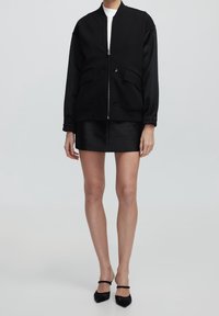 Zwarte bomberjack met een rits aan de voorkant, twee voorkruiden en geribbelde manchetten, gecombineerd met een strakke zwarte rok en spitse zwarte hakken.