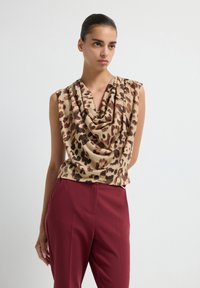 Beige mouwloze top met een gedrapeerde halslijn met een bruine luipaardprint, gecombineerd met op maat gemaakte bordeauxrode broek. Glad stoftextuur.
