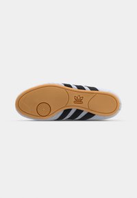 Vue de la semelle d'une basket Adidas montrant une semelle extérieure en gomme avec un motif circulaire et le logo Adidas, tige blanche avec des bandes noires partiellement visibles.