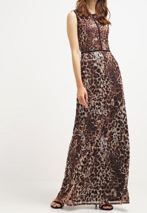 Femme porte une robe longue sans manches à imprimé léopard avec des sandales noires, debout devant un fond clair uni.