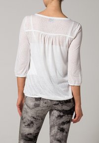 Weiße, halbtransparente Bluse mit 3/4-Ärmeln, die ein dezentes Texturmuster und einen gerafften Rücken aufweist. Kombiniert mit grauen, Batik-Jeans.