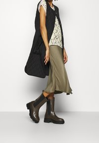 Black quilted sleeveless vest over a floral top, paired with a flowing olive skirt and brown ankle boots with black elastic sides.
