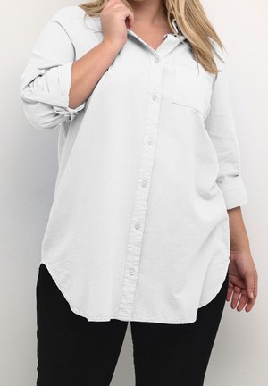 Kobieta w luźnej białej koszuli zapinanej na guziki z podwiniętymi rękawami i czarnych spodniach, stojąca na tle jednolitego jasnego tła.