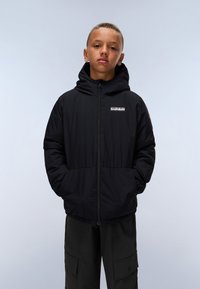 Veste puffer noire avec fermeture éclair à l'avant, capuche ajustable et poches latérales. Présente un petit logo sur la poitrine. Portée avec un pantalon ample de couleur sombre.