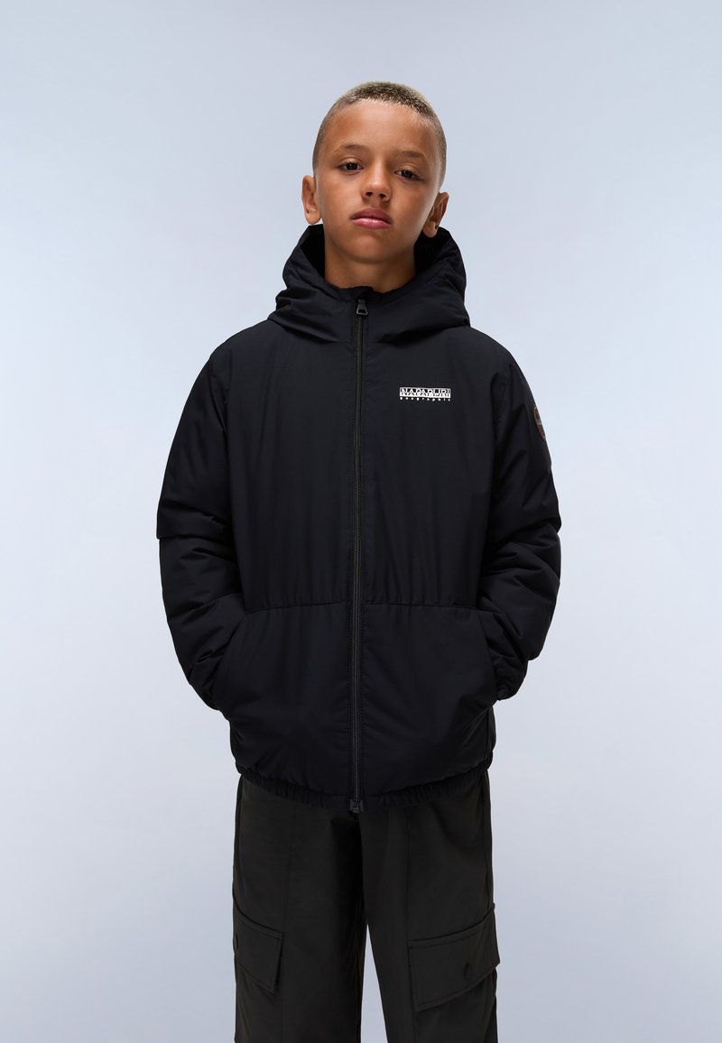 Veste puffer noire avec fermeture éclair à l'avant, capuche ajustable et poches latérales. Présente un petit logo sur la poitrine. Portée avec un pantalon ample de couleur sombre.