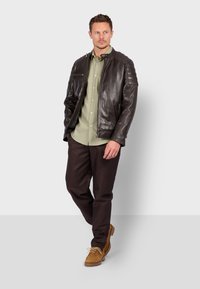 Hnědá kožená bunda se zipem vpředu, s žebrovanými detaily na ramenech, v kombinaci se světle zelenou košilí, tmavě hnědými kalhotami a béžovými botami.