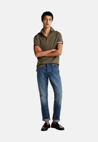 Polo verde oliva con cuello y puños a rayas, combinado con jeans azul deslavado arremangados en los bajos y mocasines marrón oscuro.
