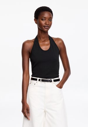 Mladá žena nosiaca čierny halter top a biele nohavice s vysokým pásom a čiernym opaskom, stojaca s jednou rukou v kapse na bielom pozadí.