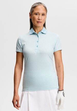 Kvinde iført en lyseblå mønstret polo, hvid nederdel og en hvid golfhandske på venstre hånd, stående foran en ensfarvet baggrund.